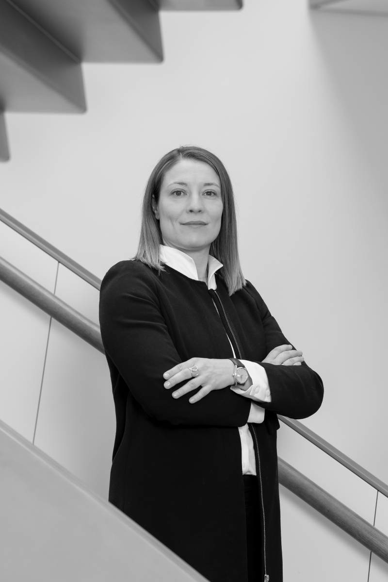 Auf dem Bild sieht man Sophie Weiß. Sie steht vor einer modernen Treppe mit klaren Linien. Sie trägt ein dunkles Jackett über einem weißen Hemd. Ihre Arme sind verschränkt, sie schaut selbstbewusst und freundlich in die Kamera. Das Bild ist in Schwarz-Weiß aufgenommen.