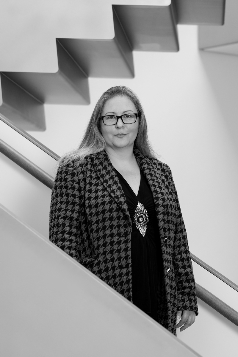 Auf dem Bild sieht man Marion Kadlec.
Sie steht vor einer modernen Treppe in der Heidi Horten Collection.
Marion trägt ein gemustertes Jackett im Hahnentritt-Muster über einem schwarzen Oberteil,
dazu ein auffälliges, großes Schmuckstück in der Mitte des Oberteils als Blickfang.
Sie hat langes, glattes Haar und trägt eine Brille.
Ihr Blick ist ruhig und freundlich, ihre Haltung sachlich und klar.
Das Foto ist in Schwarz-Weiß aufgenommen.