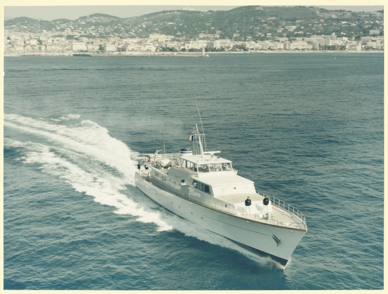 Luxusyacht fährt mit Schaumkronen durch das Wasser, im Hintergrund eine Küstenstadt und Hügel.