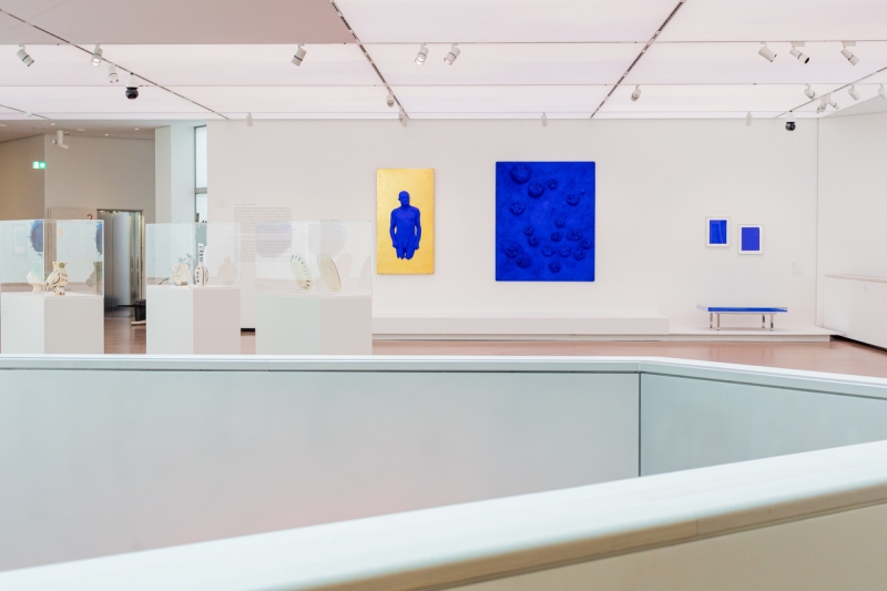Moderne Galerie mit blauen und goldenen Kunstwerken, Vitrinen mit Skulpturen und minimalistischer Sitzgelegenheit.