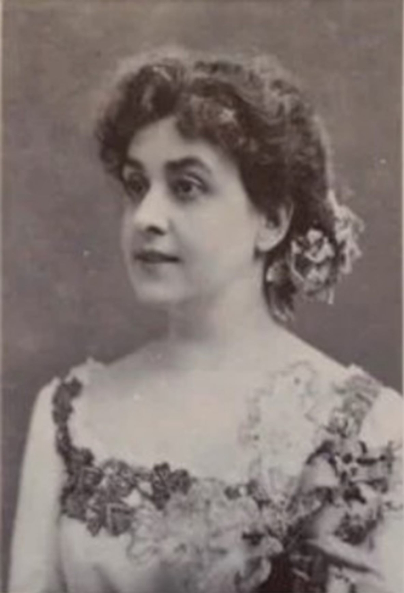 Portrait of a woman with curly hair, wearing a floral dress, gazing slightly to the side against a muted background.