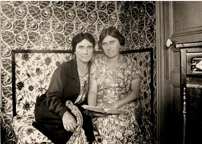 Two women sit closely together on a floral-patterned sofa, with a patterned wall behind them, holding a book.
