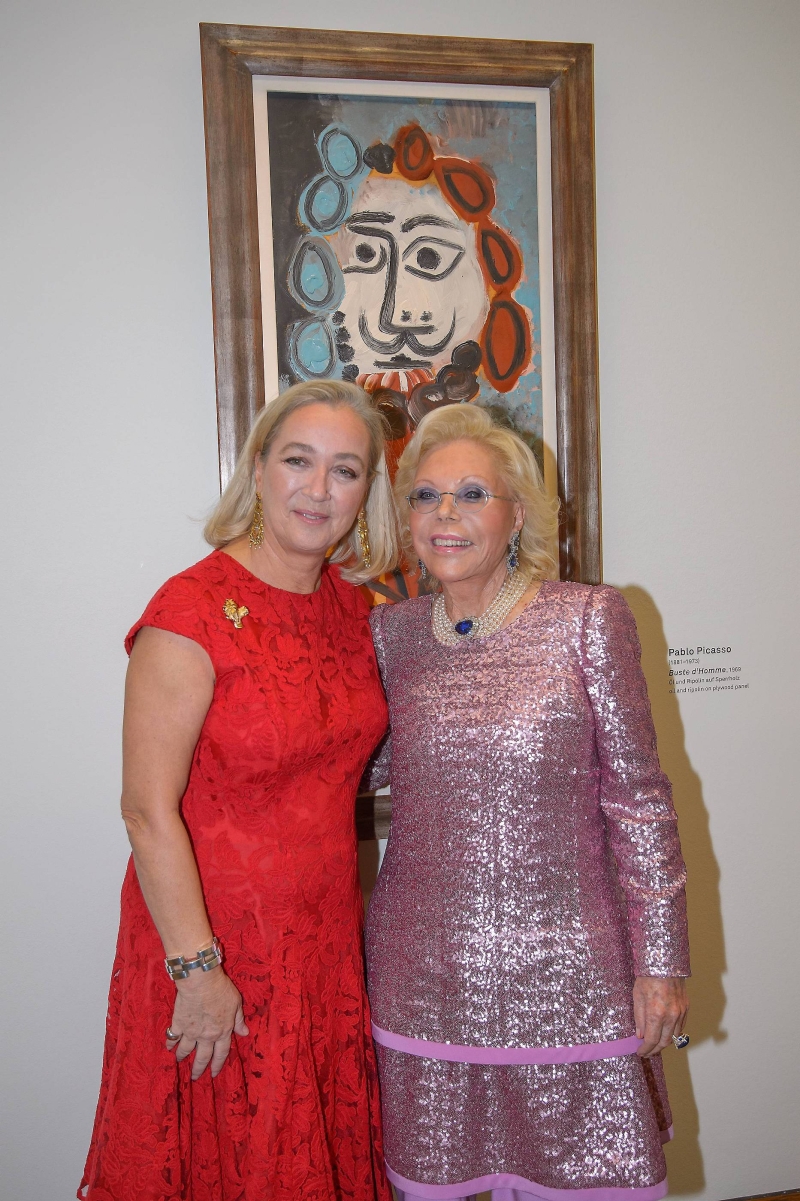 Zwei Frauen posieren lächelnd vor einem Picasso-Gemälde, eine trägt ein rotes Kleid, die andere ein glitzerndes, pinkes Outfit.