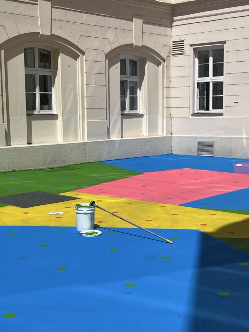 Auf dem Foto sieht man Hanuschhof in Wien. Der Boden ist in starken Farben bemalt – man erkennt große Flächen in Blau, Gelb, Grün, Pink, Schwarz und Lila.
In jeder Farbfläche sind viele kleine, bunte Punkte zu sehen. Diese Punkte zeigen, wie viel CO₂ in einem bestimmten Jahr ausgestoßen wurde – ein Punkt steht für eine Milliarde Tonnen.
Vorne in der Mitte steht ein weißer Farbeimer mit einem langen Stiel, daneben liegt ein kleines Farbtablett – man sieht, dass gerade an dem Kunstwerk gearbeitet wird oder es vor Kurzem fertiggestellt wurde.
Die Farben sind kräftig und leuchten im Sonnenlicht. Die Bemalung macht den Hof freundlich, auffällig und lebendig – und sie hat einen Zweck:
Die Spezialfarbe hilft dabei, die Hitze zu reduzieren, weil sie das Sonnenlicht besser zurückwirft als normaler Beton.
Dieses Kunstwerk heißt „CO₂ntext“. Es verbindet Umweltthema, Kunst und Stadtraum – und lädt zum Nachdenken und Verweilen ein.