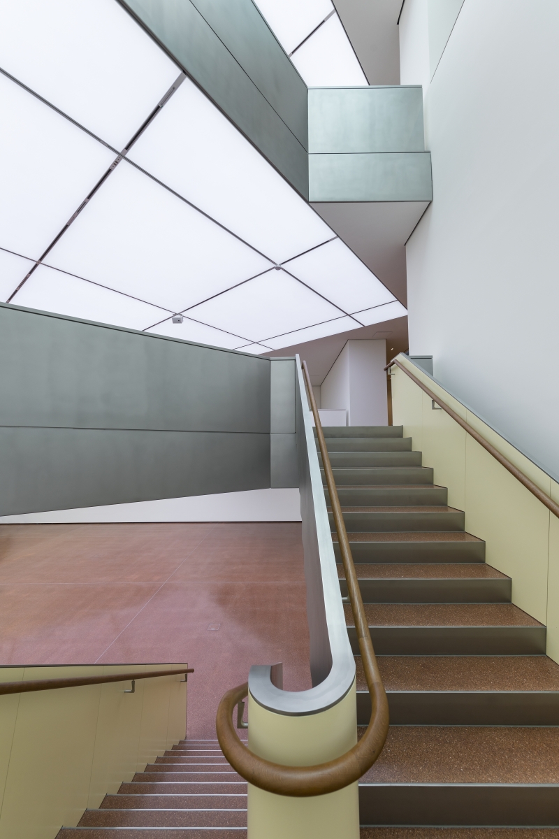 Moderne Treppe mit Holzgeländer, grauen Wänden und einem hellen, geometrischen Deckenlicht.