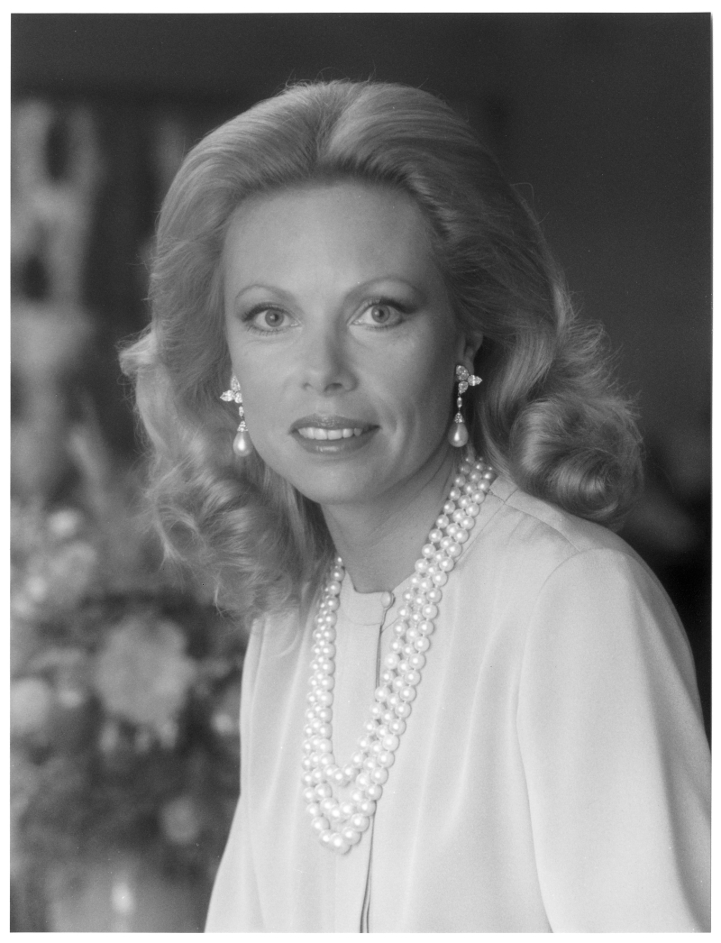 Heidi Horten with long, wavy blonde hair wears pearl necklaces and earrings, smiling softly against a blurred background.