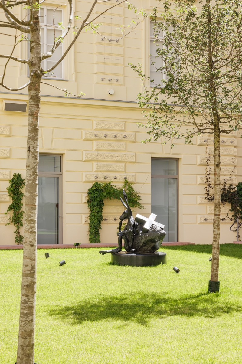 Sculpture of a seated figure with a cross on its back in a green courtyard, framed by trees and a yellow building.