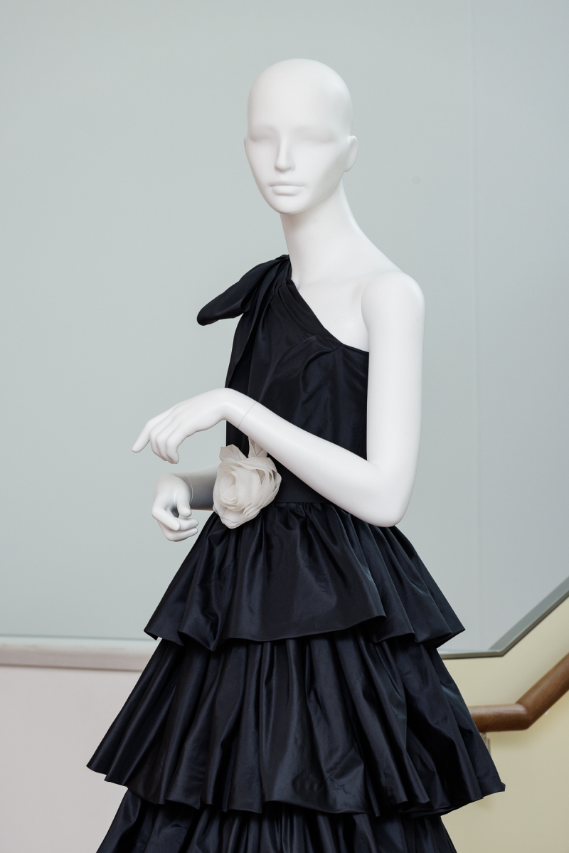 Schwarzes, schulterfreies Abendkleid mit Rüschen und einer blassrosa Blume an der Taille, präsentiert auf einer Schaufensterpuppe.