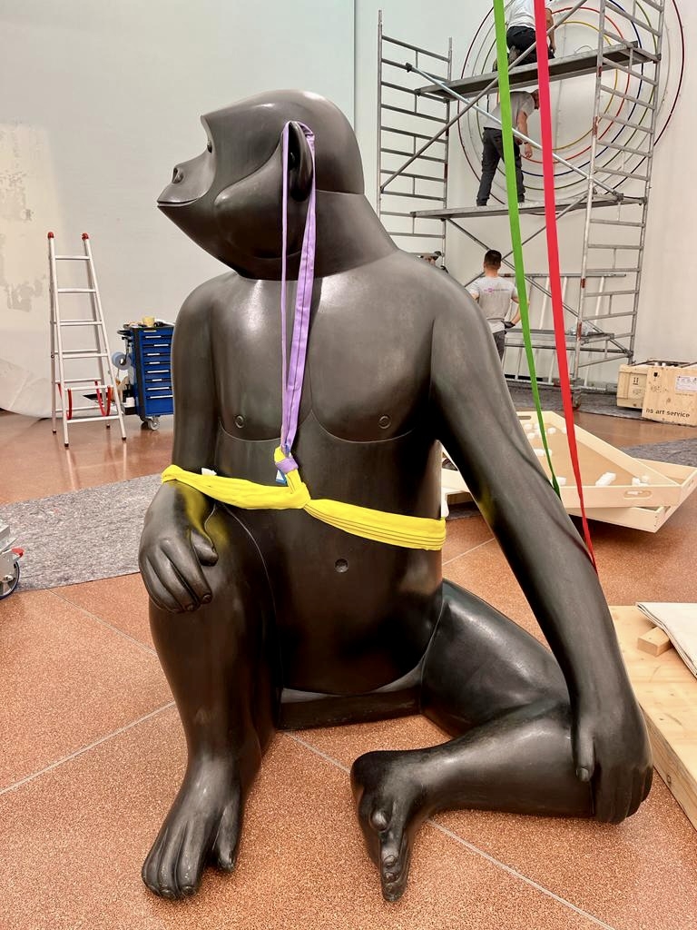 A large black gorilla sculpture sits on the floor, secured with colorful straps, in an art installation space.