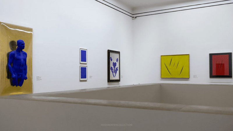 A blue sculpture stands against a white wall, surrounded by colorful abstract artworks in a gallery setting.