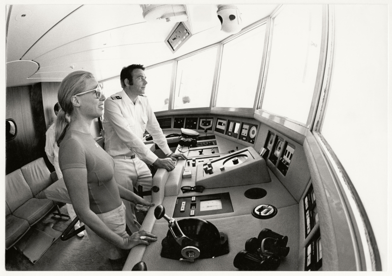 Zwei Personen stehen am Steuer eines modernen Schiffs, umgeben von Bedienfeldern und großen Fenstern.