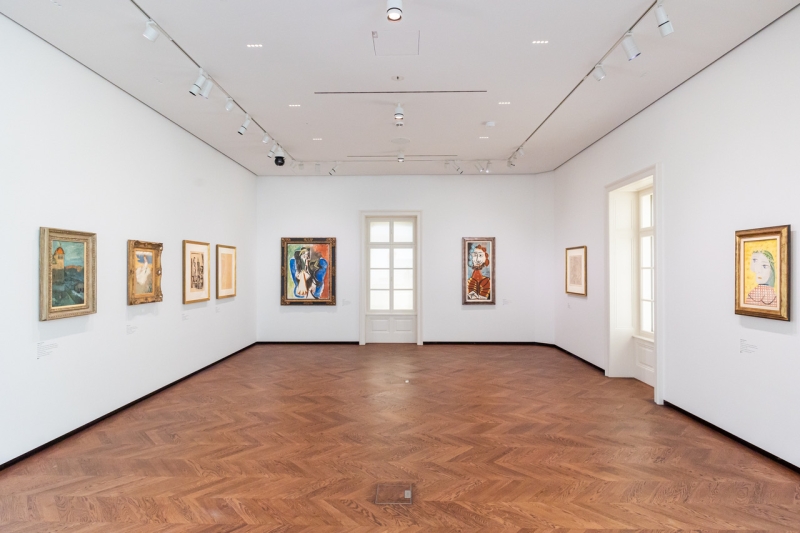 Ausstellungsraum mit mehreren Gemälden an weißen Wänden und einem Fenster mit Holzrahmen. Holzfußboden sichtbar.