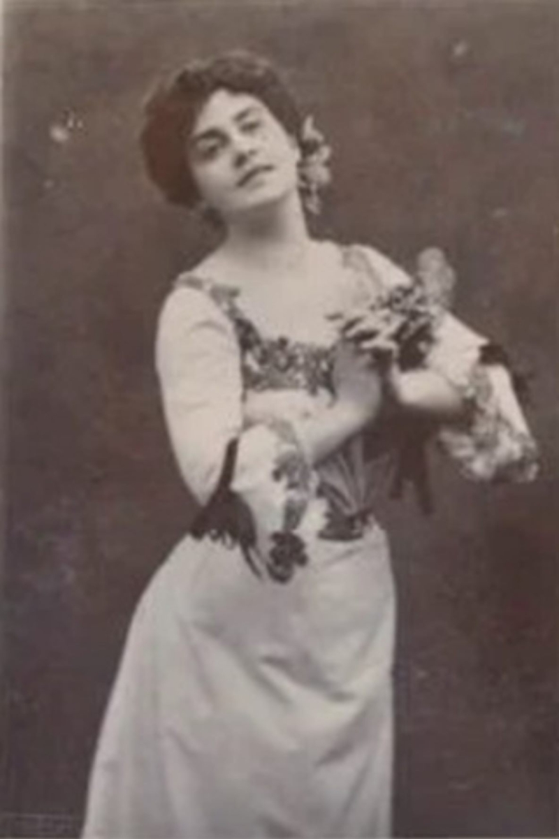 Woman in a floral dress poses gracefully, holding flowers, with a soft expression against a dark background.