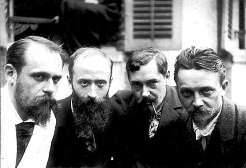 Four men with beards pose closely together, wearing formal attire, in a black-and-white photograph.