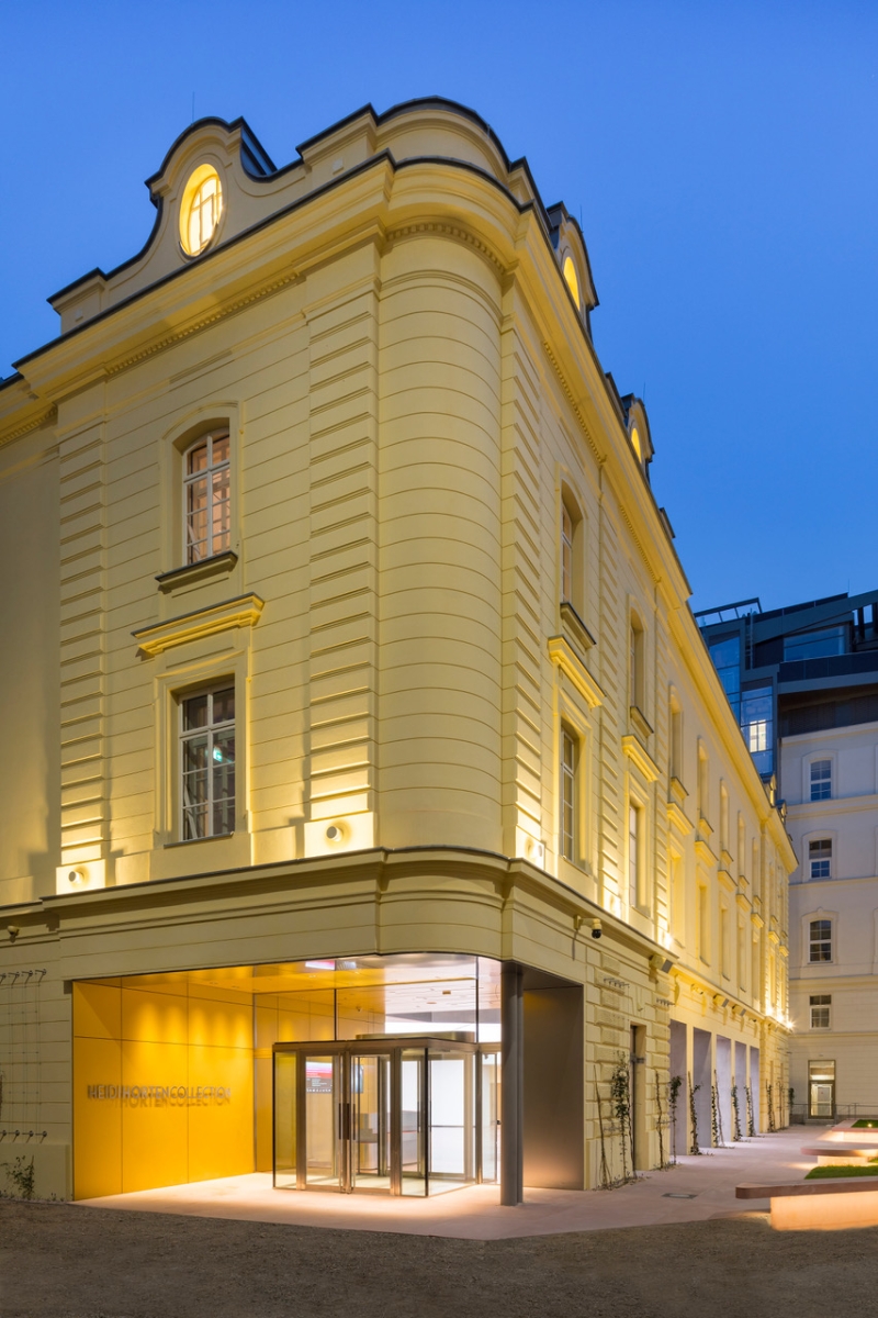 Historisches, gelbes Gebäude mit moderner Glaseingangstür, beleuchtet bei Dämmerung.