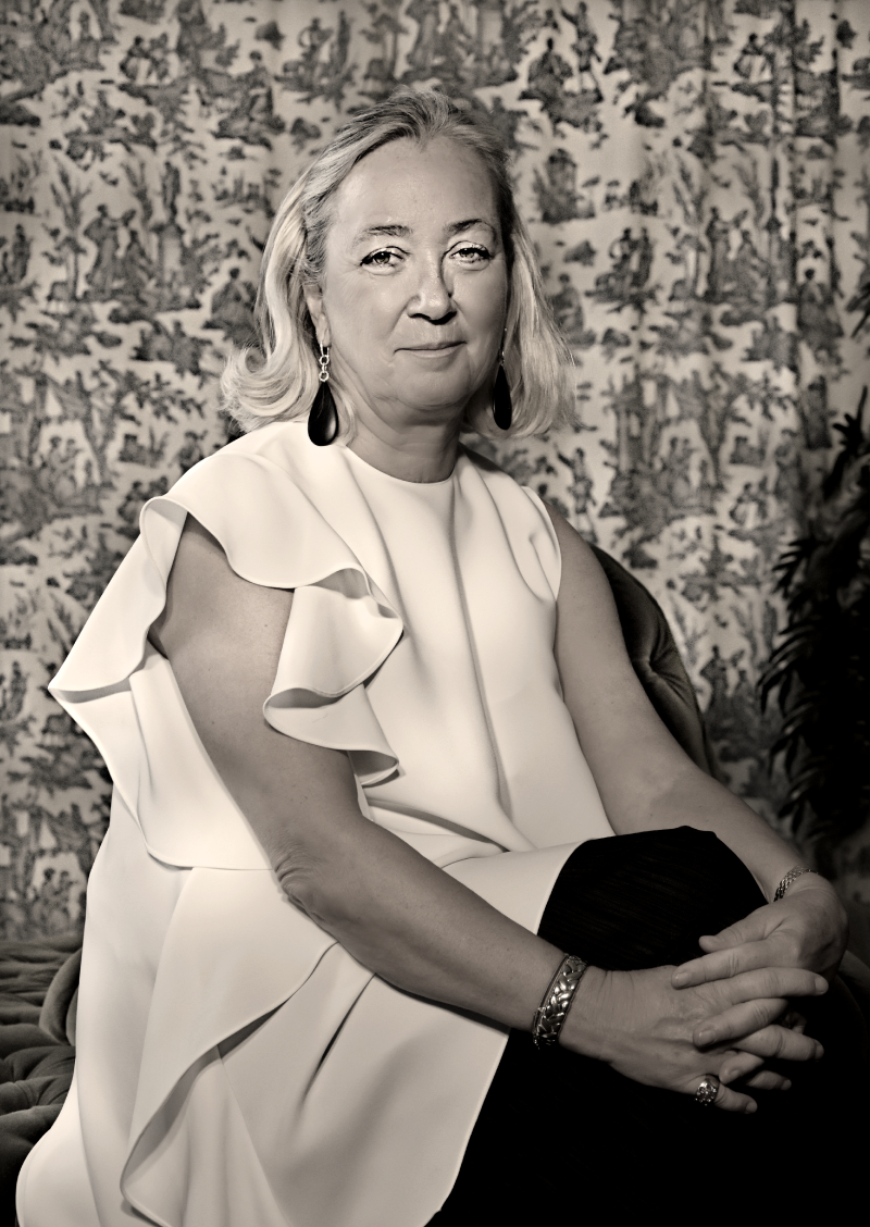 Agnes Husslein-Arco poses in a ruffled white dress, seated with hands clasped, against a patterned backdrop.