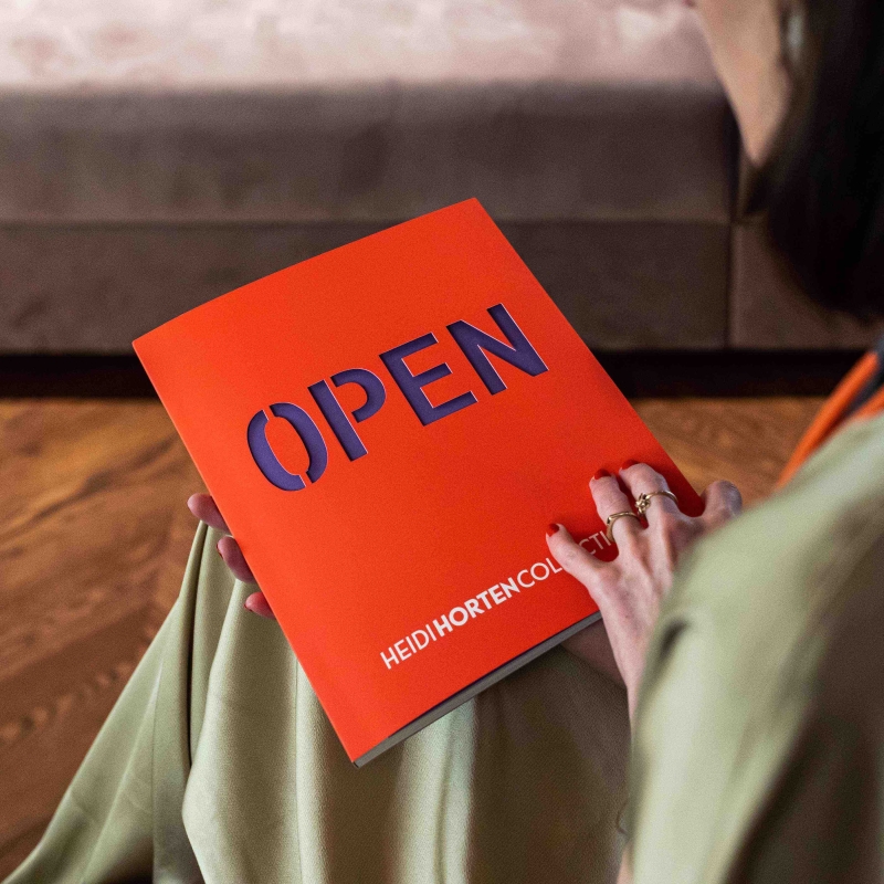 A person holds an orange booklet titled "OPEN" with dark blue text, seated on a wooden floor.