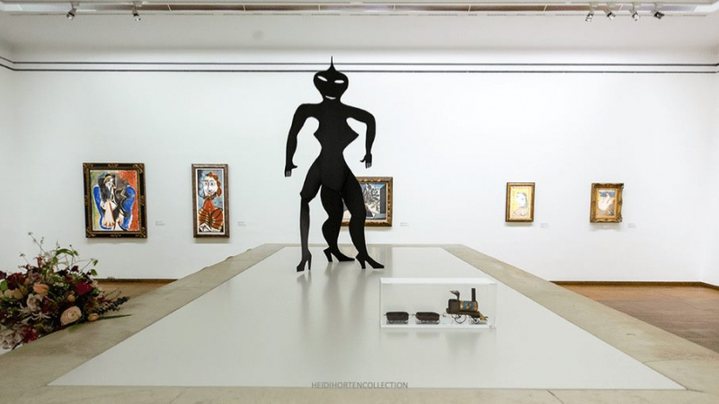 A large black sculpture stands center stage in a gallery, surrounded by colorful framed artworks on white walls.