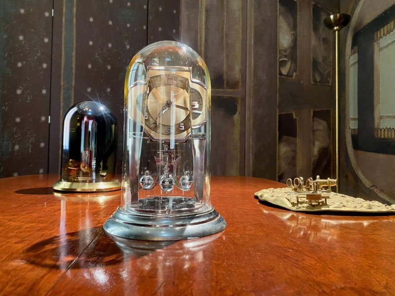 A glass dome clock with a metallic face sits on a wooden table, alongside a black dome and a decorative golden object.