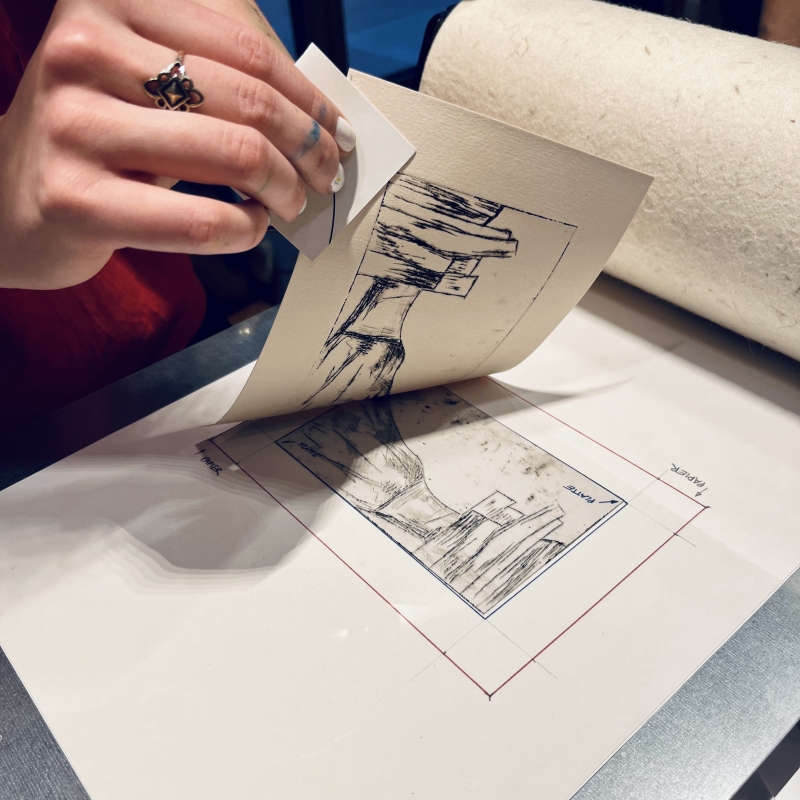 Eine Hand hebt ein Blatt Papier mit einer Zeichnung, während ein Druckwerkzeug auf einem Tisch liegt.