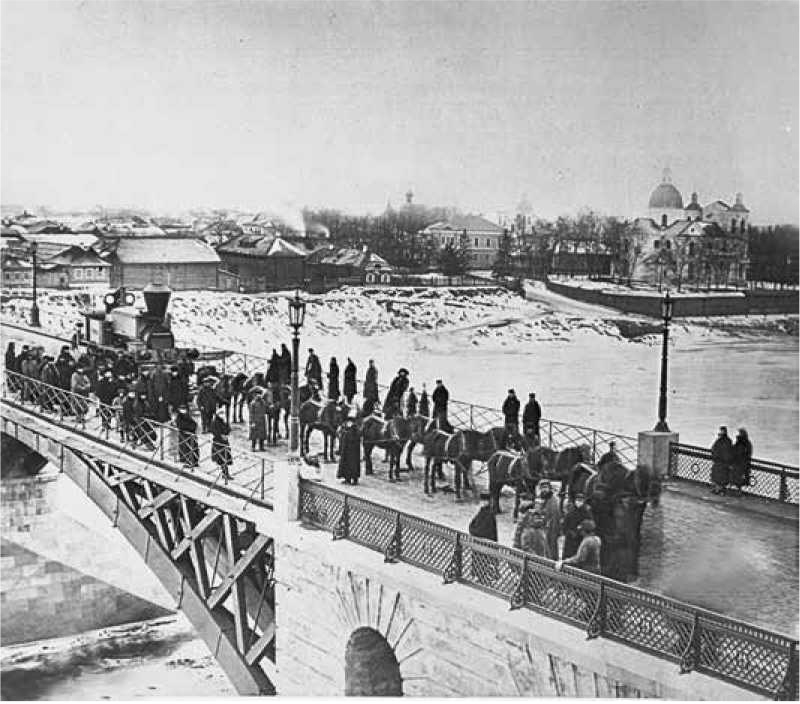 Crowd on a bridge with horses and a steam locomotive, snowy landscape and buildings in the background.