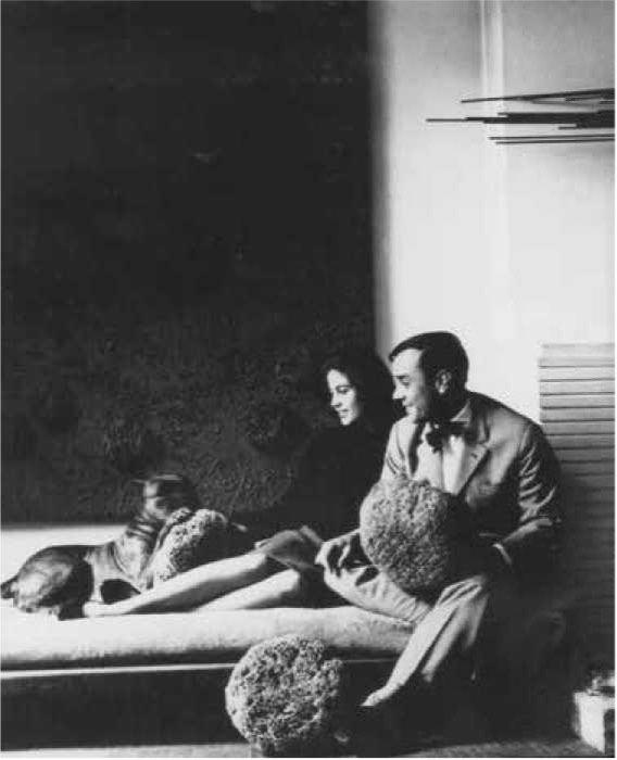 Paar sitzt auf einer Couch, umgeben von skulpturalen Objekten, mit einem Hund, vor einem dunklen Hintergrund.