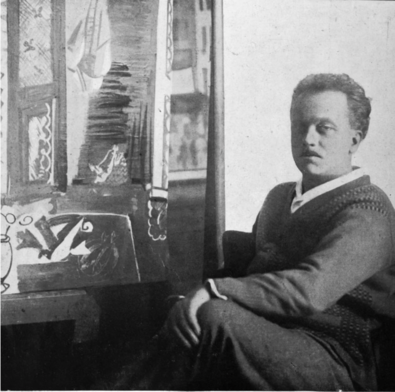 A man sits beside a colorful painting, with patterned curtains and a window in the background, exuding a contemplative demeanor.