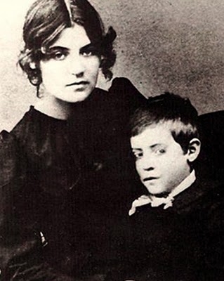A young woman with dark hair in a black dress sits closely with a boy in a dark outfit, both looking serious.