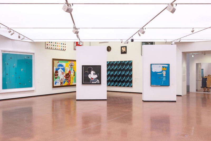 Art gallery interior featuring colorful paintings, white walls, and polished floors under bright overhead lighting.