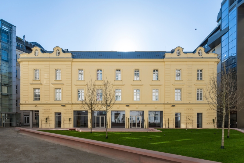 Historisches, gelbes Gebäude mit großen Fenstern, umgeben von modernem Glasbau und grünem Rasen.