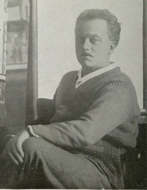 Raoul Dufy mit kurzem Haar, in einem Pullover sitzend, Blick zur Seite, im Hintergrund unscharfe Kunstwerke.