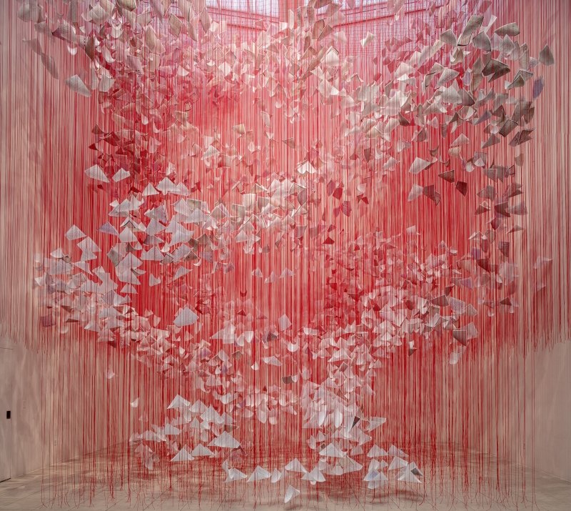 Die Installation füllt einen Ausstellungsraum mit einem dichten Geflecht aus leuchtend roten Fäden, die von der Decke bis zum Boden gespannt sind. Innerhalb dieses Netzes schweben hunderte weißer Papierbögen – sie wirken wie gefaltete Briefe, lose Blätter oder kleine Flügel. Die Briefe breiten sich wie in einer Luftbewegung spiralförmig nach oben oder außen auszubreiten. Die rote Farbe der Fäden dominiert den Raum. Die Fäden sind so zahlreich und dicht, dass sie aus der Ferne wie ein schimmernder Vorhang wirken, der die Grenzen des Raums verändert und ein begehbares, fast körperliches Erlebnis erzeugt.