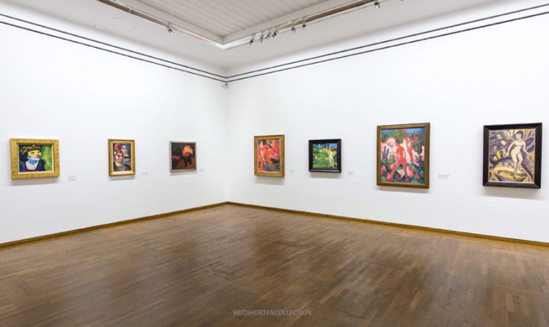 Gallery interior featuring eight colorful paintings on white walls, with wooden flooring and overhead lighting.