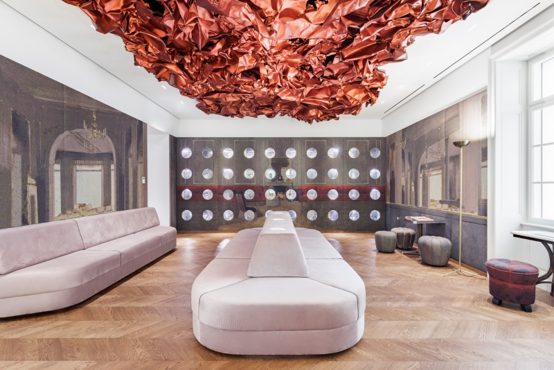 Auf dem Foto sehen wir den Tea Room im Museum der Heidi Horten Collection in Wien.
An der Decke hängt ein großes, rotes Kunstwerk von Hans Kupelwieser.
Es sieht aus wie zerknittertes Papier oder glänzender Stoff,
wurde aber aus Aluminium gemacht.
An der hinteren Wand gibt es viele runde Fenster – sie sehen aus wie Bullaugen.
Dahinter befinden sind kleine, kostbare Objekte aus der Sammlung von Heidi Horten.
Diese Vitrinenwand wurde von Markus Schinwald entworfen.
Auch die rosa Sitzmöbel in der Mitte des Raums gehören zu seinem Konzept.
Sie laden ein, sich hinzusetzen, zu schauen und zu entspannen.
Die Wände sind mit großen Stoffbildern bedeckt,
die an ein altes Café oder Schlosszimmer erinnern.