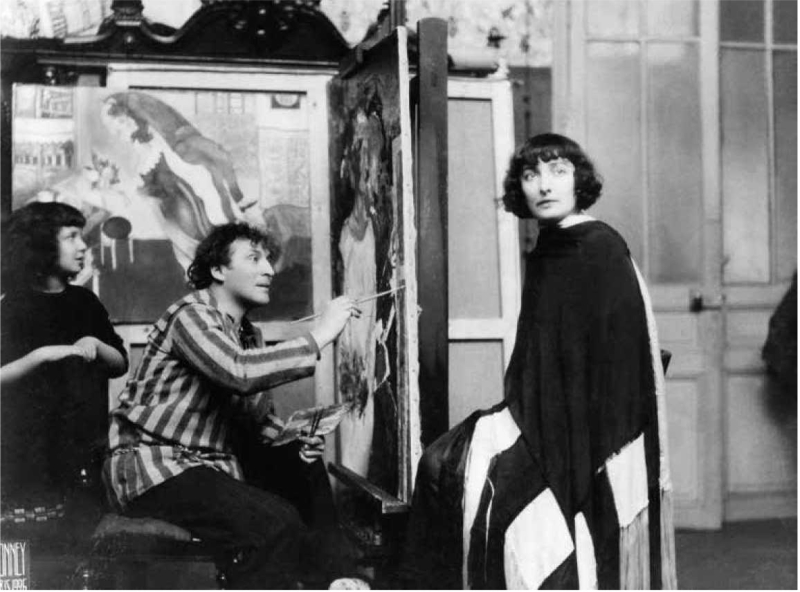 Artist in striped shirt paints a woman in a black and white cloak, with a girl observing in a studio filled with artworks.