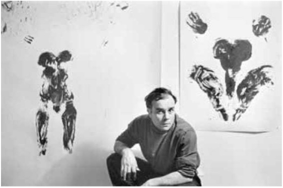 Mann in grauem Pullover sitzt vor zwei abstrakten Schwarz-Weiß-Gemälden an einer Wand.