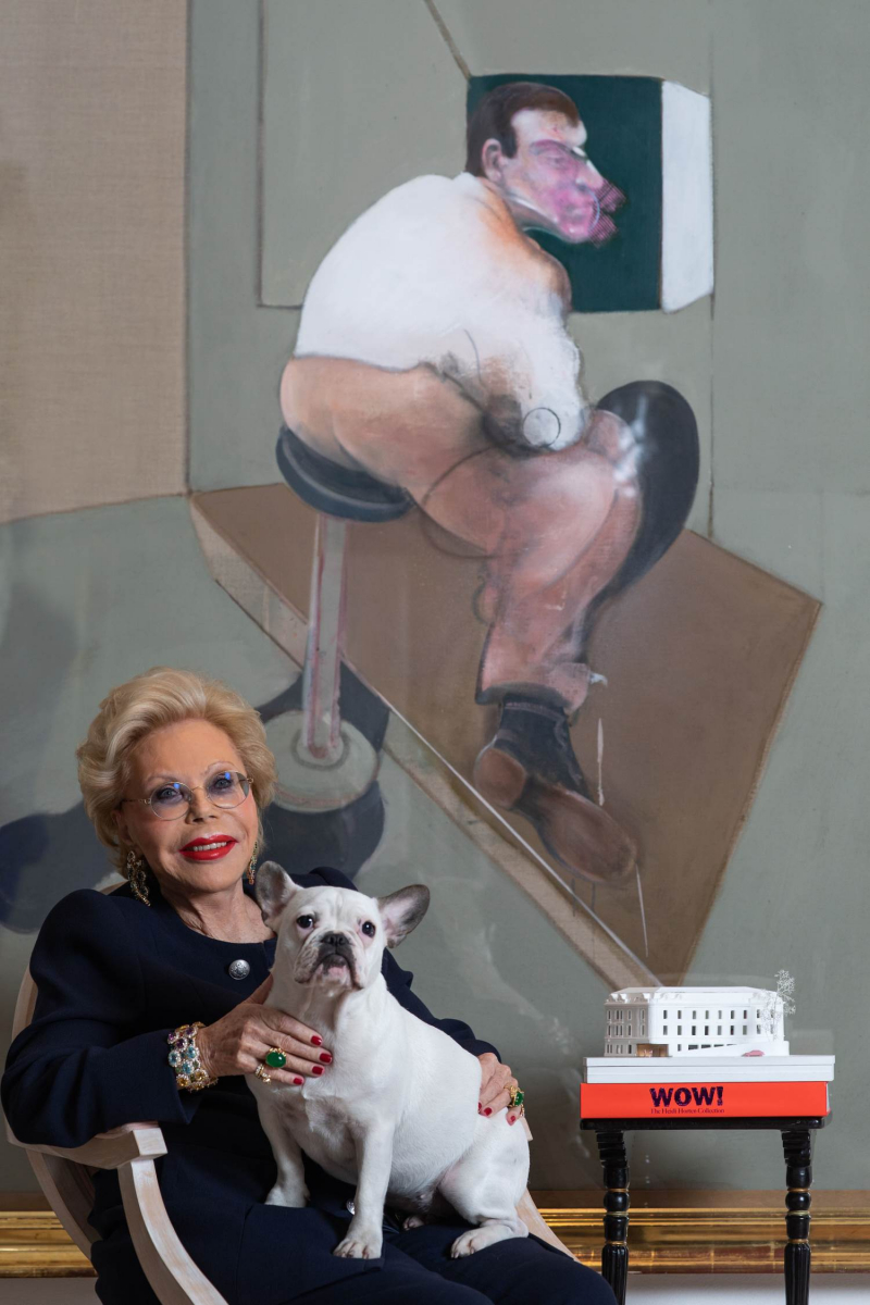 Auf dem Foto sieht man Heidi Horten.
Sie sitzt auf einem Sessel und hält einen kleinen weißen Hund auf dem Schoß.
Heidi Horten trägt dunkle Kleidung, große Schmuckstücke und eine blaue Brille.
Sie lächelt freundlich in die Kamera.
Hinter ihr hängt ein großes Gemälde von Francis Bacon.
Das Bild zeigt eine sitzende Figur, die sehr ausdrucksstark und ungewöhnlich gemalt ist – typisch für Bacons Stil.
Neben Heidi Horten steht ein kleiner Tisch.
Darauf liegen Kataloge, z.B. der "WOW" Katalog der Heidi Horten Collection.
Oben auf dem Tisch sieht man ein weißes Modell des Museumsgebäudes.