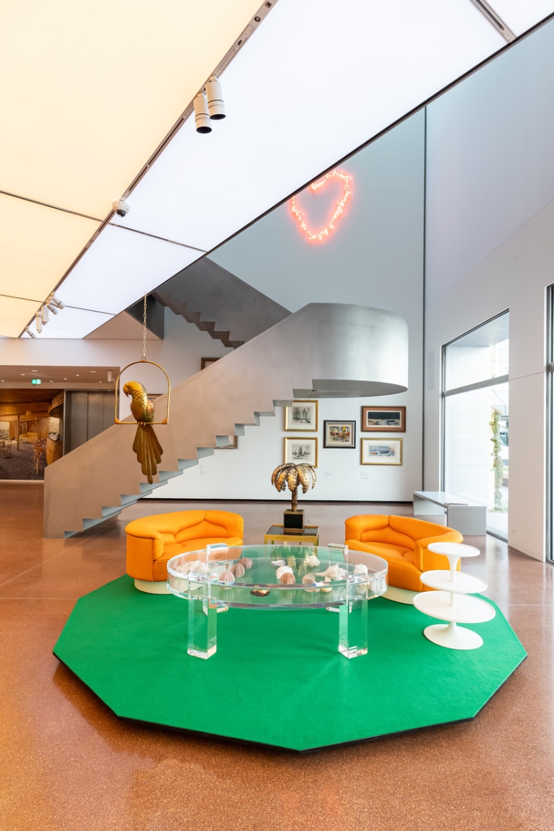 Moderne Lounge mit orangefarbenen Sesseln, gläsernem Tisch, grüner Teppich und einer Treppe im Hintergrund.