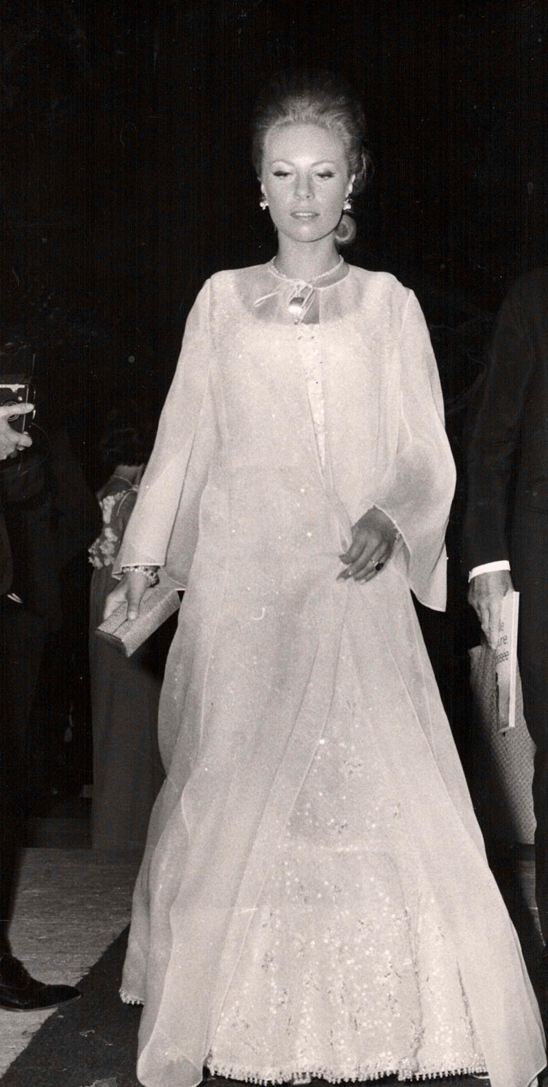 Frau in eleganter, weißer Abendgarderobe mit langen Ärmeln, umgeben von Menschen und Kameras, 1960er Jahre.