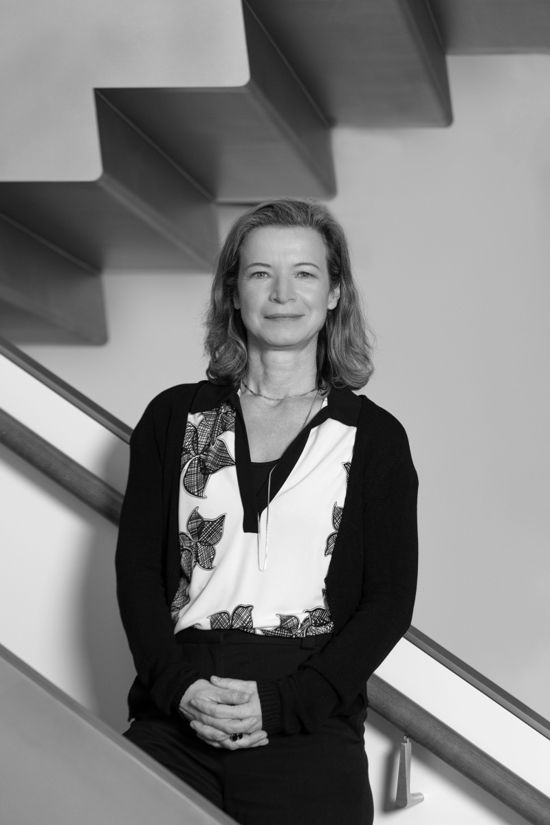 Auf dem Bild sieht man Bettina Hagen.
Sie steht vor einer modernen Treppe mit klaren Linien.
Bettina trägt eine weiße Bluse mit schwarzem Blumenmuster
und darüber eine schlichte schwarze Strickjacke.
Ihre Hände sind ruhig gefaltet, und sie schaut freundlich und offen in die Kamera.
Das Foto ist in Schwarz-Weiß aufgenommen.