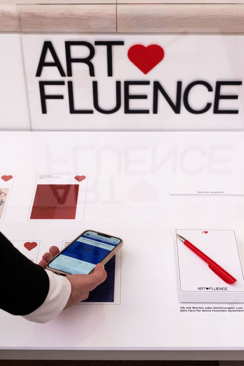 Hand mit Smartphone über einer Tischoberfläche mit Karten und einem roten Stift, im Hintergrund "ART ♥ FLUENCE".