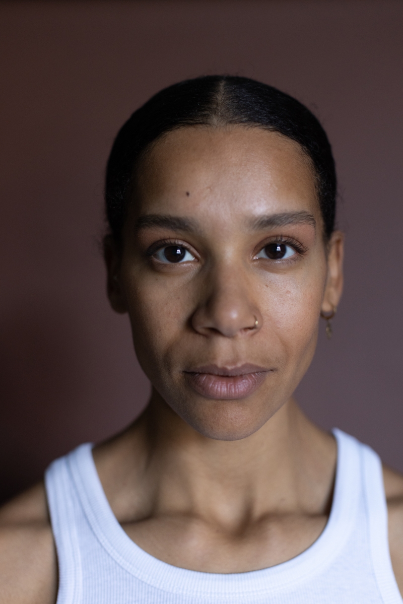 Porträt einer Frau mit dunklen Haaren, braunen Augen und einem schlichten weißen Oberteil, vor einem rosa Hintergrund.