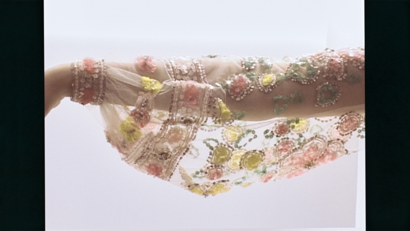 Durchsichtiger Stoff mit bunten, handgestickten Blumenmotiven in Rosa, Gelb und Grün an einem Arm.