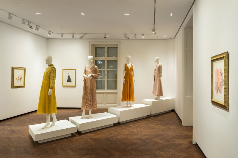 Modeausstellung mit vier Schaufensterpuppen in eleganten Kleidern, umgeben von Kunstwerken an der Wand.