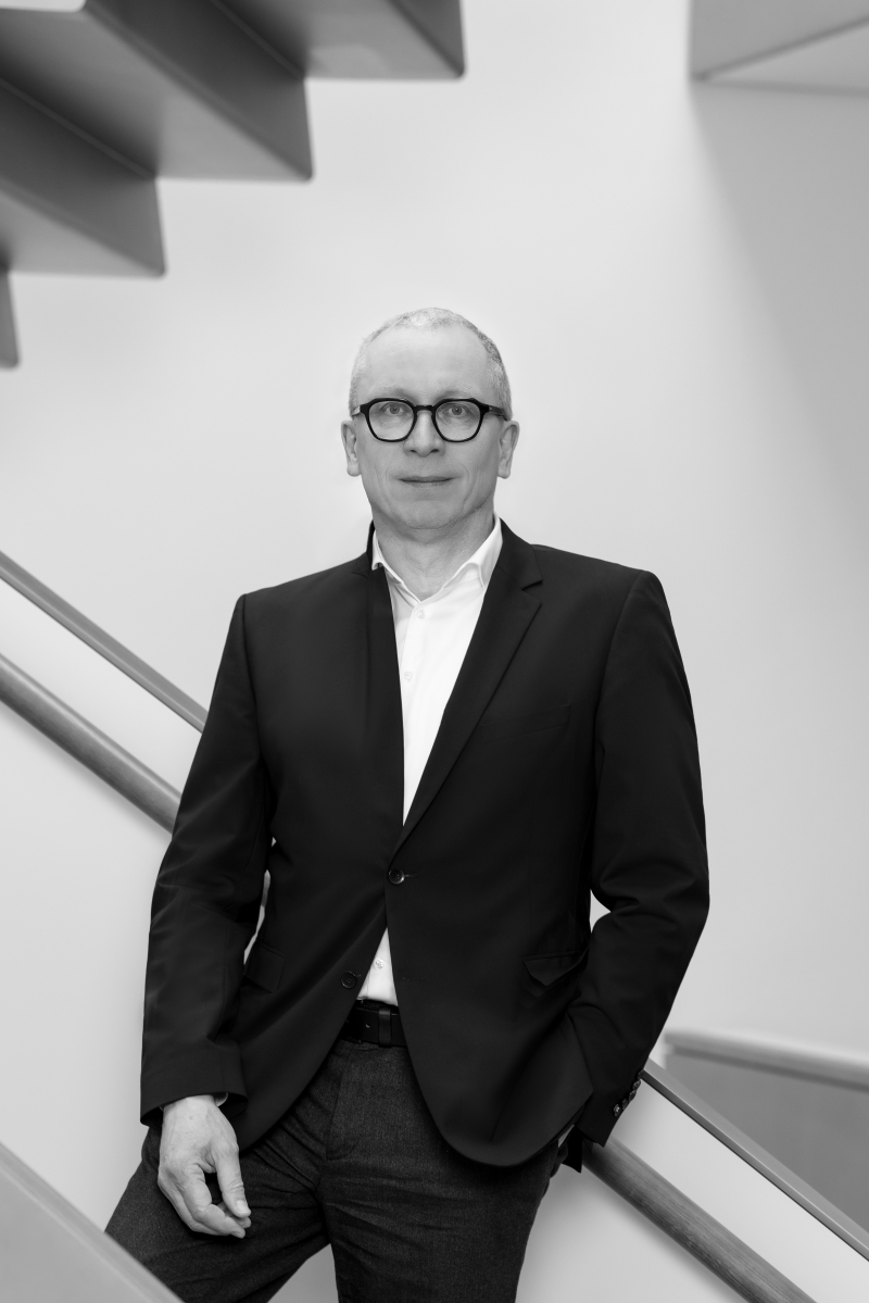 Auf dem Bild sieht man Rolf H. Johannsen.
Er steht vor einer modernen Treppe mit klaren Linien.
Er trägt einen dunklen Anzug, ein weißes Hemd und eine Brille mit rundem Rahmen.
Seine Hände sind locker, eine steckt in der Hosentasche,
und er blickt ruhig und freundlich in die Kamera.
Das Foto ist in Schwarz-Weiß aufgenommen.