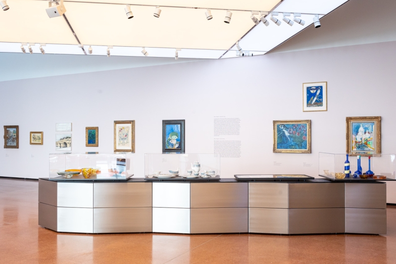 Moderne Galerie mit Gemälden an der Wand und einem Ausstellungsstück aus Glas und Metall in der Mitte.