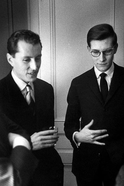 Two men in formal attire, one holding a drink, engaged in conversation against a light-colored background.