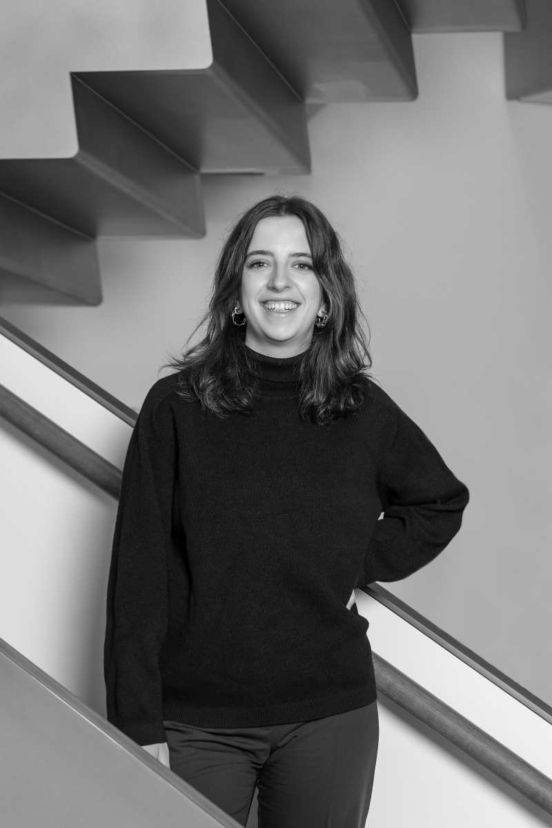 Auf dem Bild sieht man Annkathrin Weber.
Sie steht vor einer modernen Treppe in der Heidi Horten Collection.
Annkathrin trägt einen dunklen Rollkragenpullover und eine dunkle Hose.
Ihr Outfit ist schlicht und modern.
Sie hat mittellanges, gewelltes Haar und trägt runde Ohrringe.
Sie blickt offen und freundlich lächelnd in die Kamera.
Eine Hand stützt sie locker in die Hüfte.
Das Foto ist in Schwarz-Weiß aufgenommen.