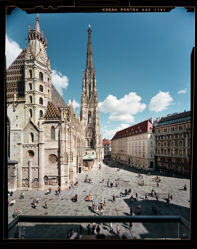 Auf dem Foto sehen wir den Stephansdom in Wien.
Das Bild wurde von einem Balkon aus gemacht.
Der Stephansdom ist eine sehr alte Kirche.
In der Mitte sieht man einen sehr hohen, spitzen Turm.
Das Dach hat bunte Ziegel. Es sieht schön und besonders aus.
Links ist die Kirche, rechts sind andere Häuser.
Auf dem Platz davor sind viele Menschen.
Der Himmel ist blau. Es gibt ein paar weiße Wolken.
Das Foto wurde mit einer alten Kamera gemacht, die einen Film benutzt.
Der Fotograf heißt Stefan Oláh.
Die Farben im Bild sind warm und angenehm.