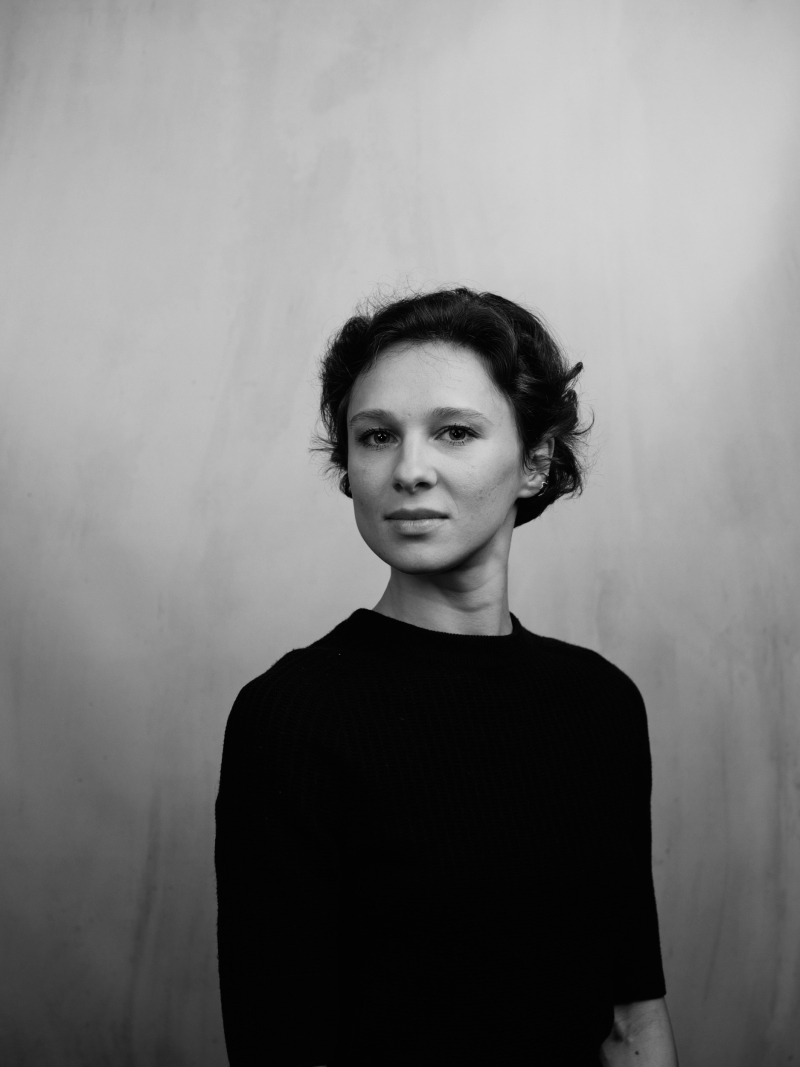 Frau mit kurzem, gewelltem Haar in schwarzem Pullover, vor einer neutralen, hellen Wand.